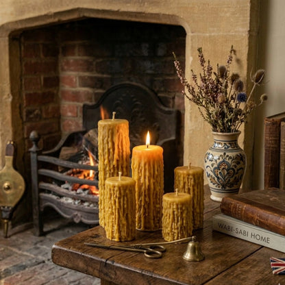 A set of four graduating-height beeswax pillar candles with a deep drip texture. One is clearly lit in the front. The candles are displayed on pages from an old book, showing their detailed, golden-yellow texture against a dark fabric background.