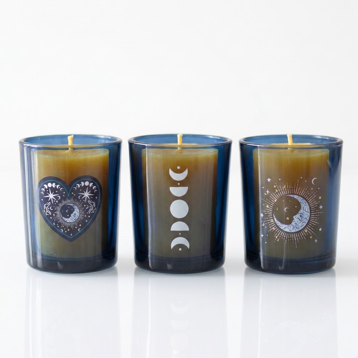 A set of three cobalt blue glass votive candle holders with distinct silver celestial designs including moon phases and stars, holding unlit wicks on a white reflective surface.