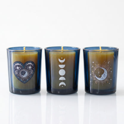 A set of three cobalt blue glass votive candle holders with distinct silver celestial designs including moon phases and stars, holding unlit wicks on a white reflective surface.