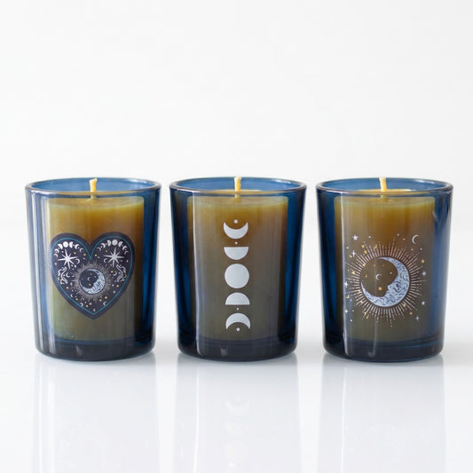A set of three cobalt blue glass votive candle holders with distinct silver celestial designs including moon phases and stars, holding unlit wicks on a white reflective surface.