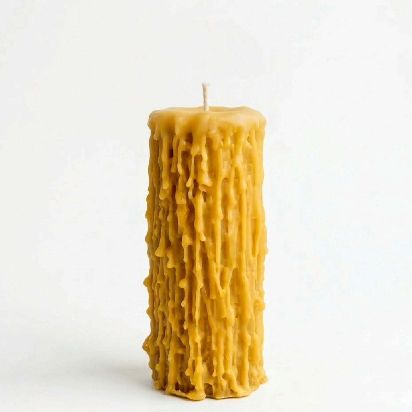 A high-angle photograph showing a group of beeswax pillar candles with distinct drip textures arranged on a rustic wooden table with pages from old books. In the background, there is a jar with dried poppy pods and other stems, and a grey fabric, creating a warm lifestyle scene.