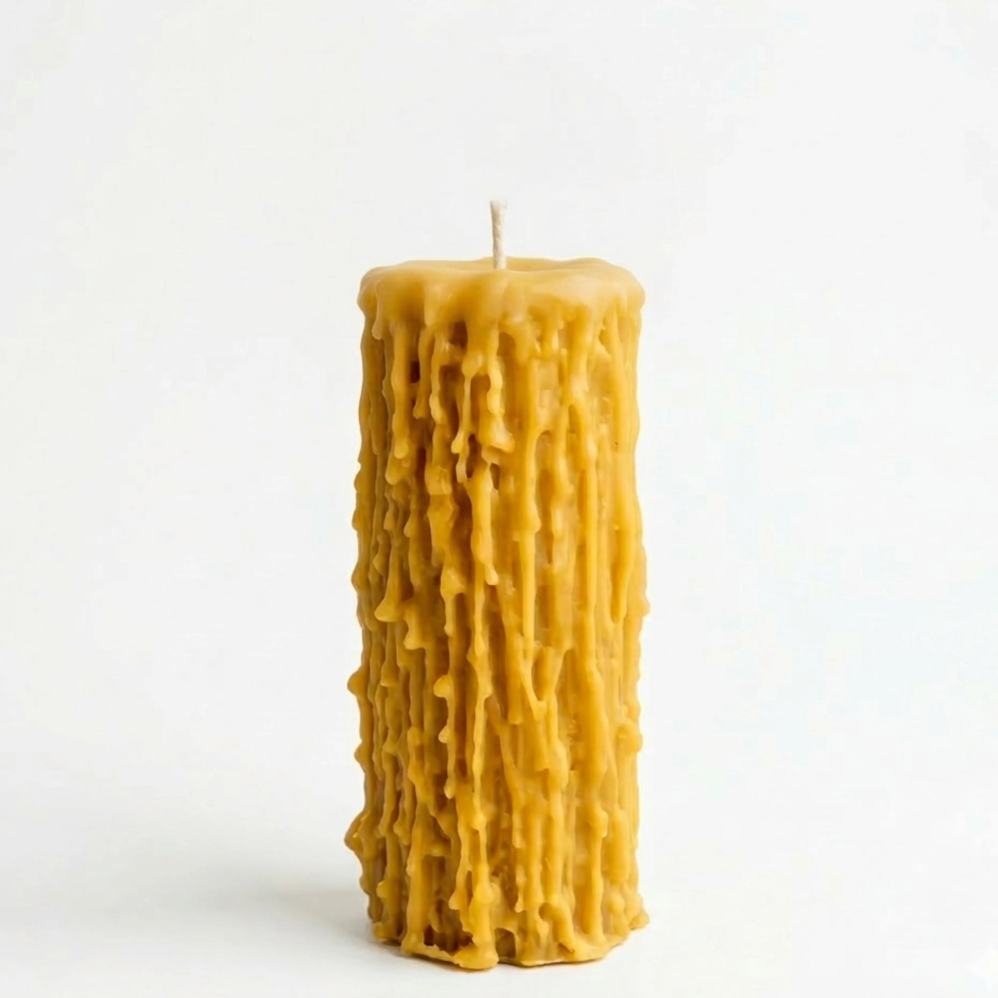 A high-angle photograph showing a group of beeswax pillar candles with distinct drip textures arranged on a rustic wooden table with pages from old books. In the background, there is a jar with dried poppy pods and other stems, and a grey fabric, creating a warm lifestyle scene.