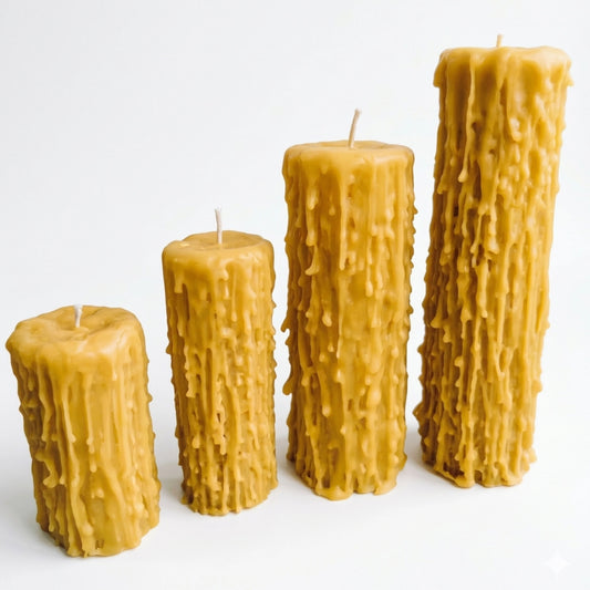 A high-angle close-up view looking down on a group of beeswax candles. The central candle is in focus, showing its top surface, cotton wick, and detailed drip texture. Other candles are out of focus in the background, along with blurred book pages and a wooden surface.