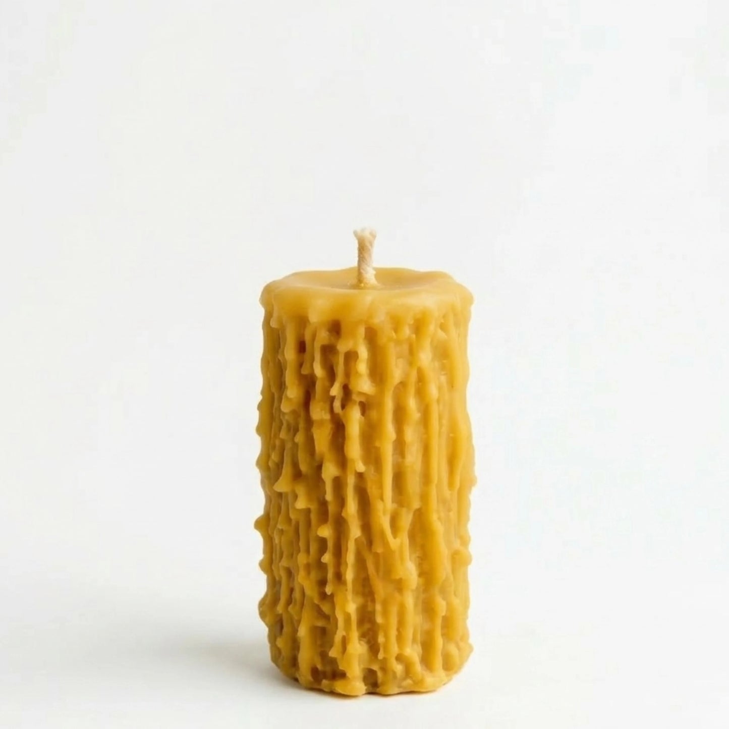 A single, lit ochre-yellow beeswax pillar candle with a natural, heavily dripped, textured surface stands on a rustic grey wooden table. Behind it, a blur of dried botanical stems and pages from an old book create a cozy scene